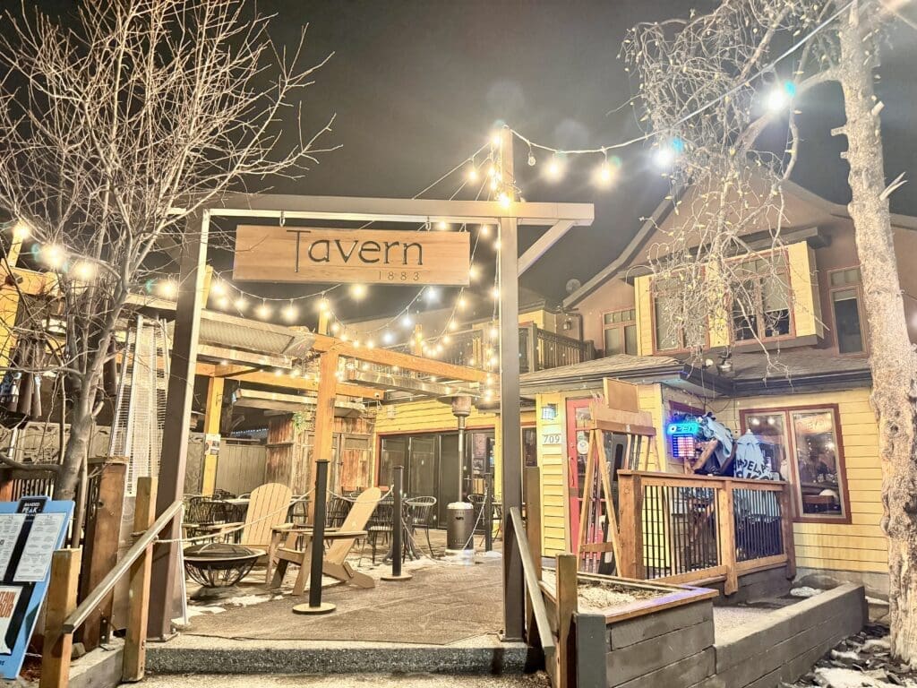 Cozy outdoor patio at Tavern 1883 in Canmore, Alberta, illuminated with string lights on a winter night. The rustic restaurant features Adirondack chairs, wooden decor, and a welcoming atmosphere perfect for evening dining and drinks in the Canadian Rockies.