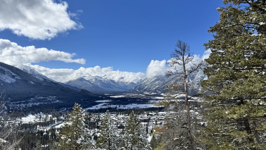 Panoramic winter view from a higher scenic point on Tunnel Mountain Trail in Banff, Alberta, overlooking snow-covered trees, the town of Banff, and the surrounding Canadian Rockies under a blue sky with scattered clouds.