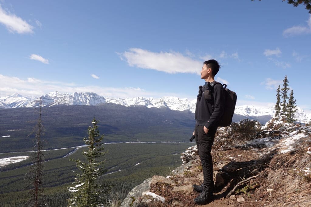 Hiker standing on a rocky ledge along Castle Lookout Trail in Banff, admiring the expansive view of the snow-covered Canadian Rockies under a bright blue sky.