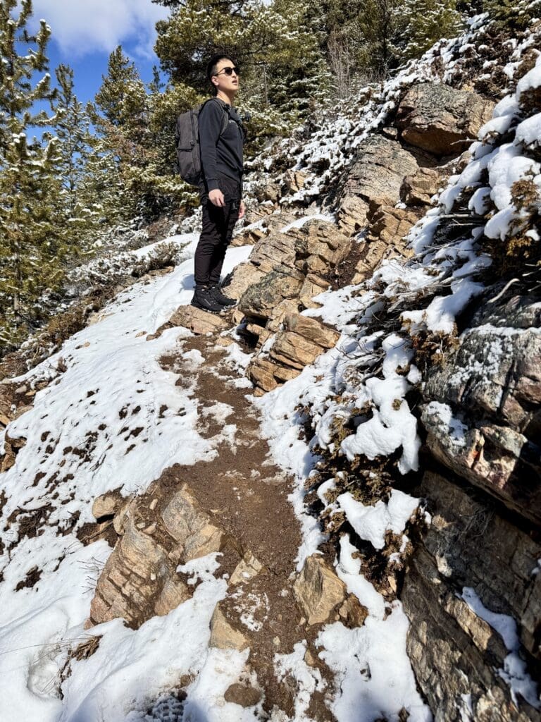 Hiker navigating a rugged and partially snow-covered rocky path where the Castle Lookout Trail in Banff becomes indistinct, surrounded by coniferous forest and alpine terrain.