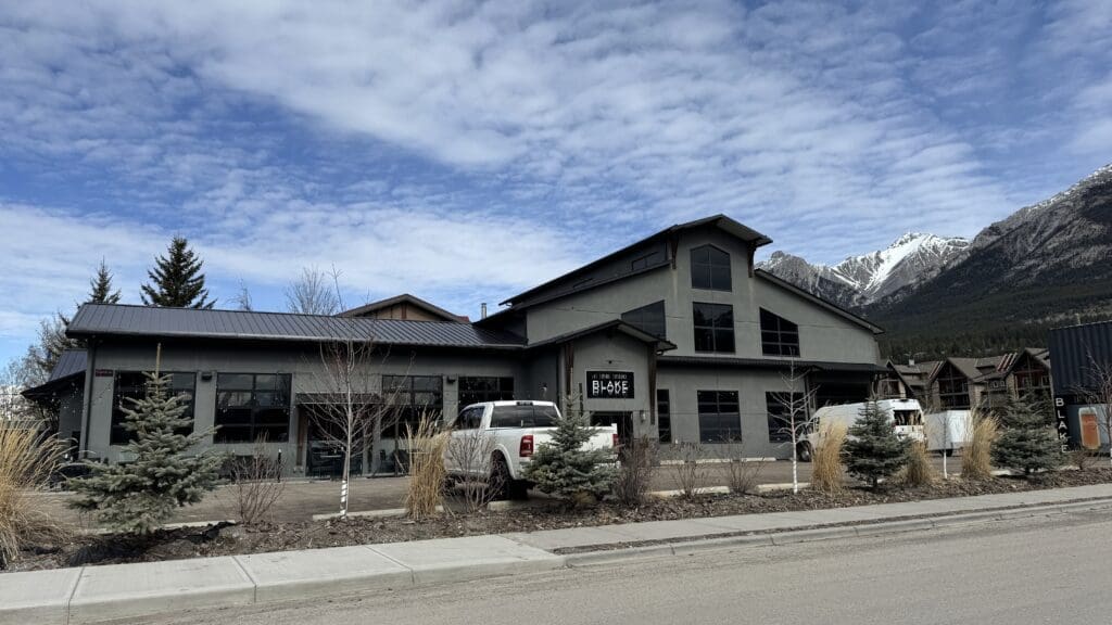 Exterior of BLAKE Restaurant Brewhouse and Distillery in Canmore, Alberta, featuring a modern industrial design with mountain views and parked vehicles out front under a partly cloudy sky.