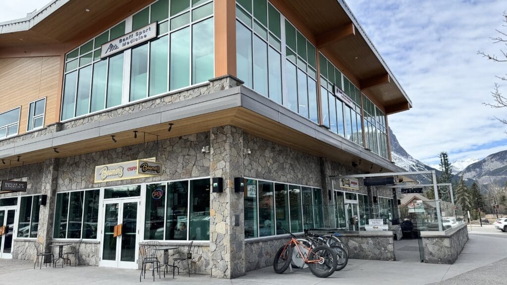 Exterior of Beamer’s Coffee Bar in Canmore, located in a modern stone and wood building with large glass windows, patio seating, and mountain views in the background under a partly cloudy sky. The best Restaurants to Dine in Canmore