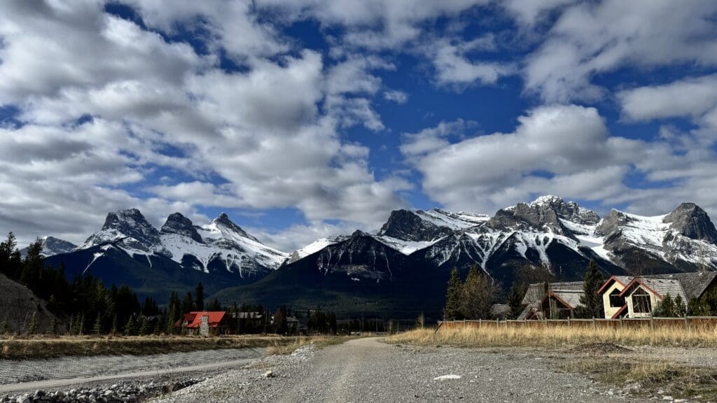 Stunning panoramic view of snow-capped Three Sisters Mountains from Cougar Creek Trailhead in Canmore, Alberta, with rustic cabins and dramatic clouds under a vibrant blue sky.