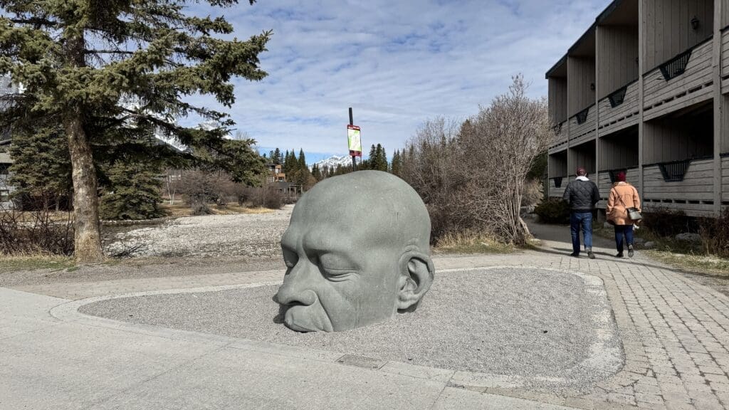 Big Head sculpture in downtown Canmore, Alberta – a large stone face partially buried in gravel with mountains in the background and pedestrians nearby