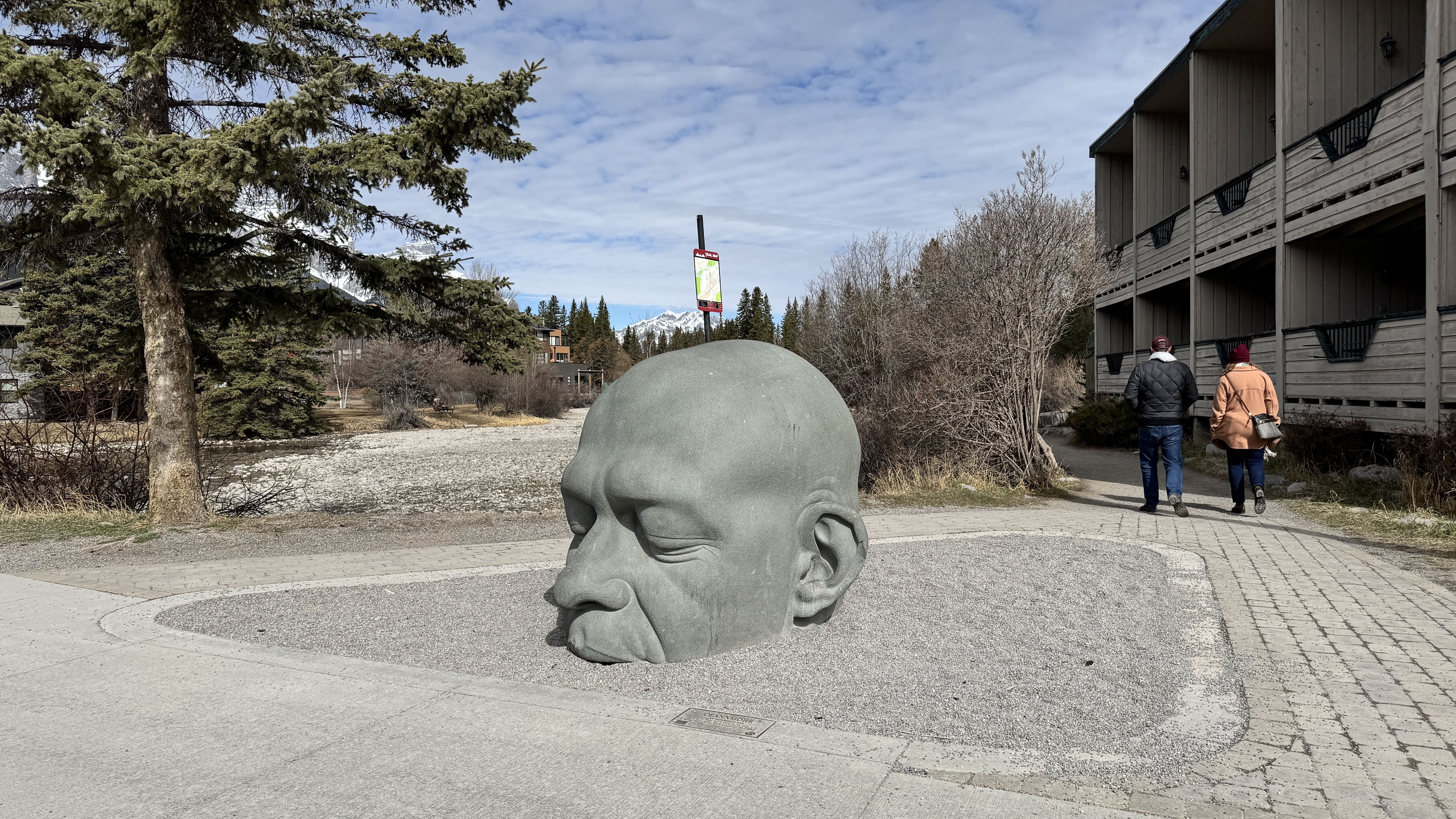 Big Head sculpture in downtown Canmore, Alberta – a large stone face partially buried in gravel with mountains in the background and pedestrians nearby