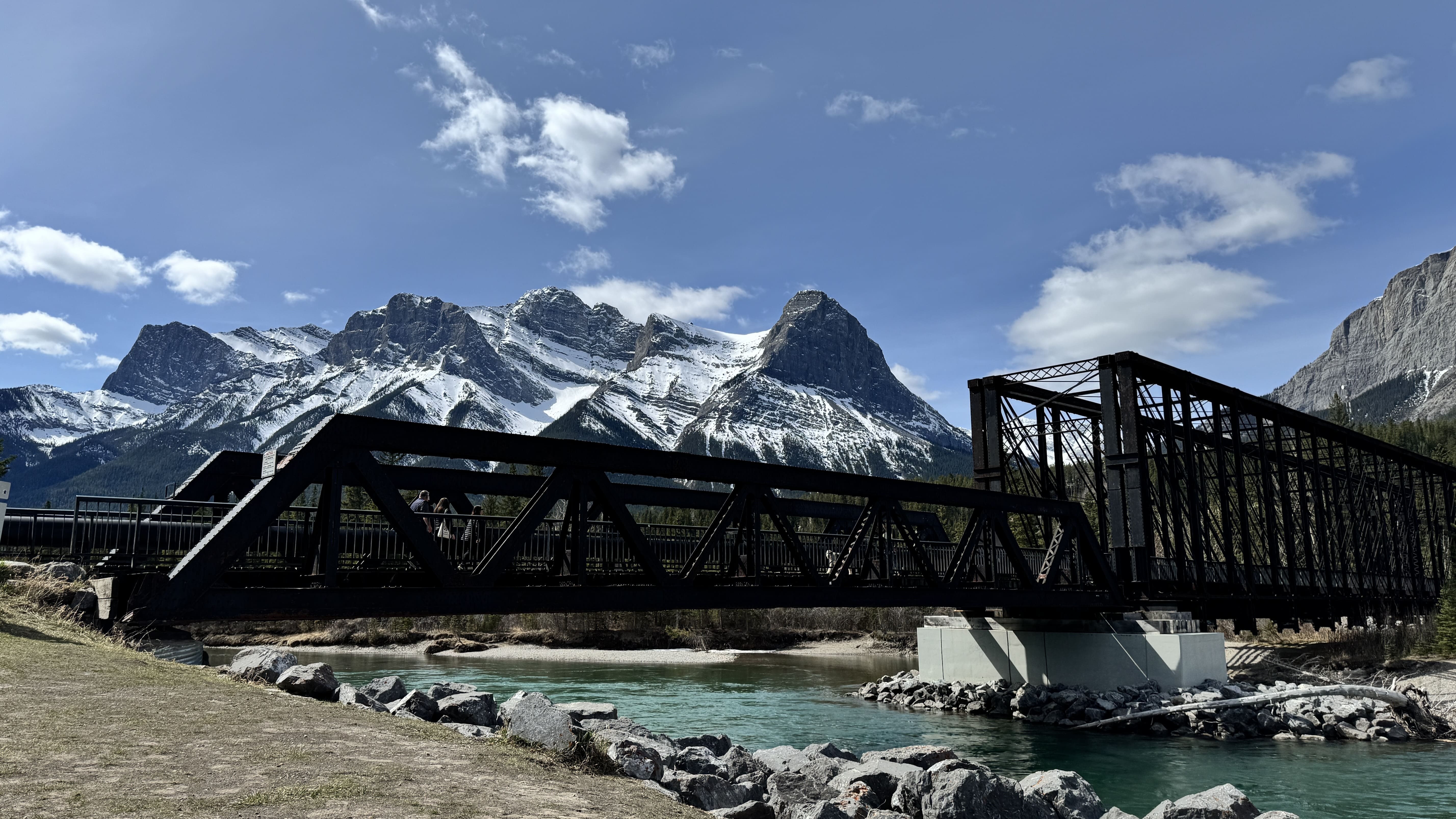 Historic Engine Bridge spanning the turquoise Bow River in Canmore, Alberta, with a backdrop of the snow-capped Rocky Mountains under a clear blue sky.