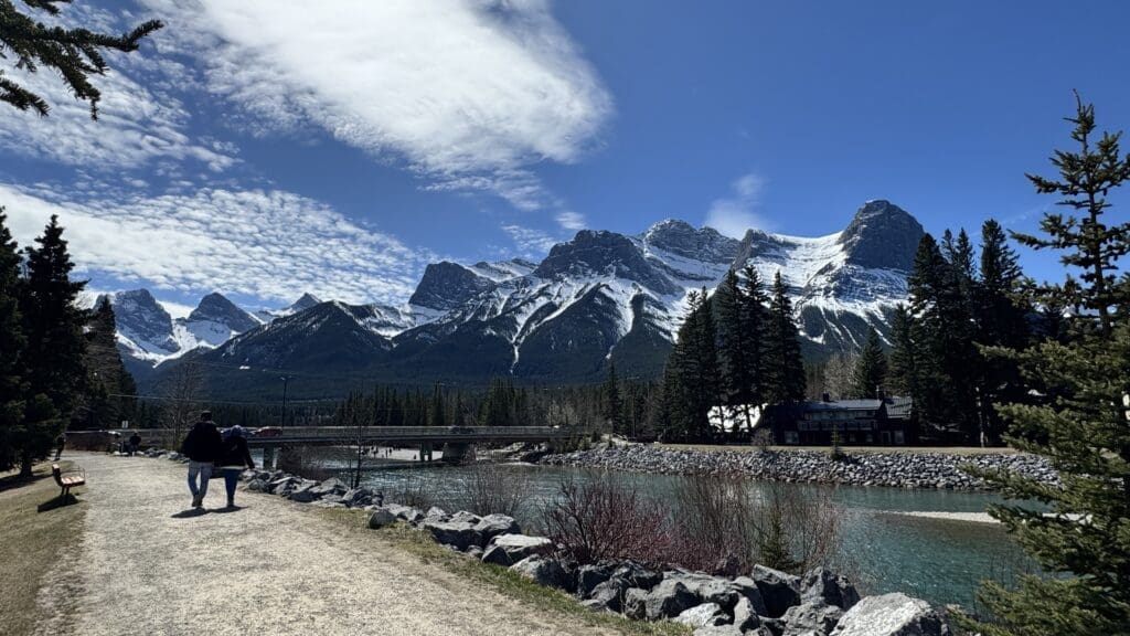 Scenic walking path along Bow River in Canmore, Alberta, featuring Canmore’s Bridge Road crossing the river, with towering Rocky Mountains in the background under a bright blue sky.