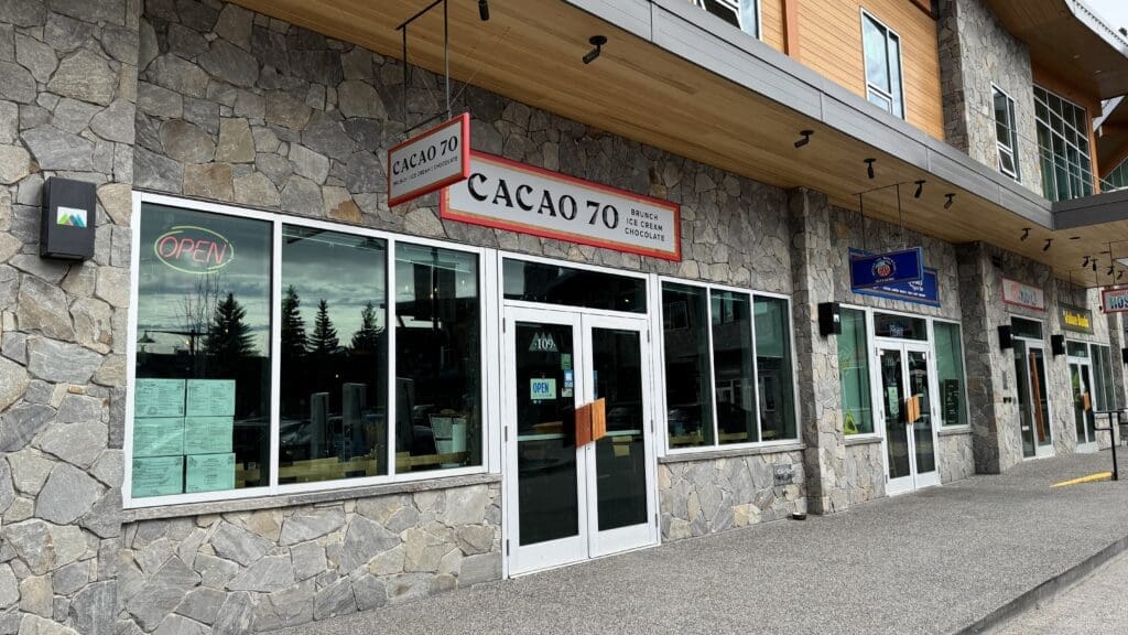 Front view of Cacao 70 in Canmore, Alberta, showcasing the stone façade, large front windows, and a prominent red-and-white sign above the entrance indicating brunch, ice cream, and chocolate offerings
