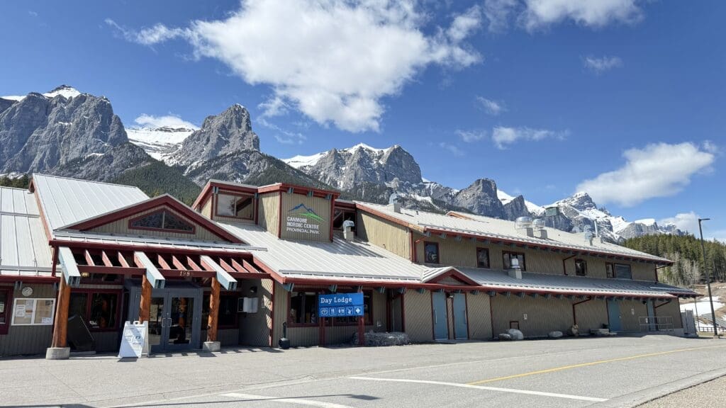 Front view of the Canmore Nordic Centre Day Lodge set against the dramatic backdrop of the Rocky Mountains and snow-covered Three Sisters peaks in Alberta, Canada.