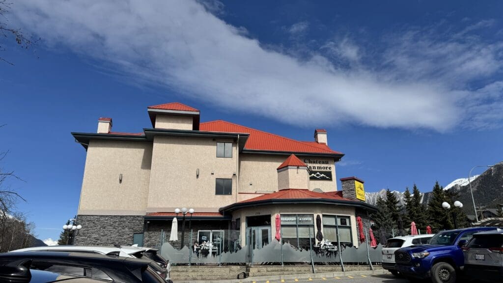 Exterior view of Chez Francois Restaurant and Patio at Chateau Canmore, featuring a beige and stone building with red roofs, outdoor seating with mountain-themed frosted glass dividers, and a backdrop of the Canadian Rockies under a vibrant blue sky. The best Restaurants to Dine in Canmore