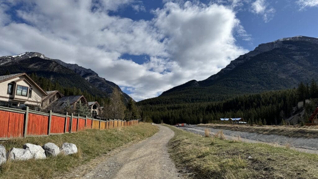 Scenic view along Cougar Creek Trail in Canmore, Alberta, showing winding gravel path beside red fences, rustic mountain homes, and dense evergreen forest under a partly cloudy sky with rugged mountain slopes in the background.