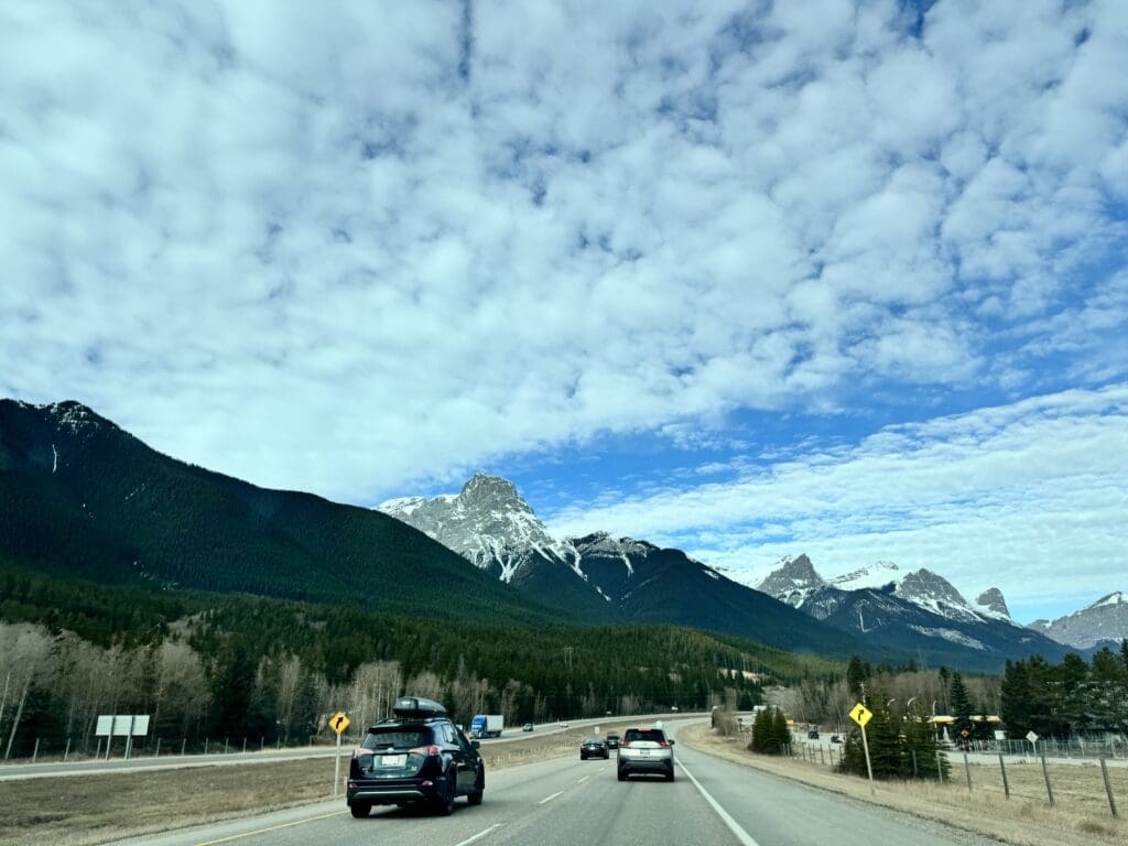 Cars driving toward Canmore Town on the Trans-Canada Highway with a panoramic view of the snow-dusted Rocky Mountains under a dramatic cloud-filled sky