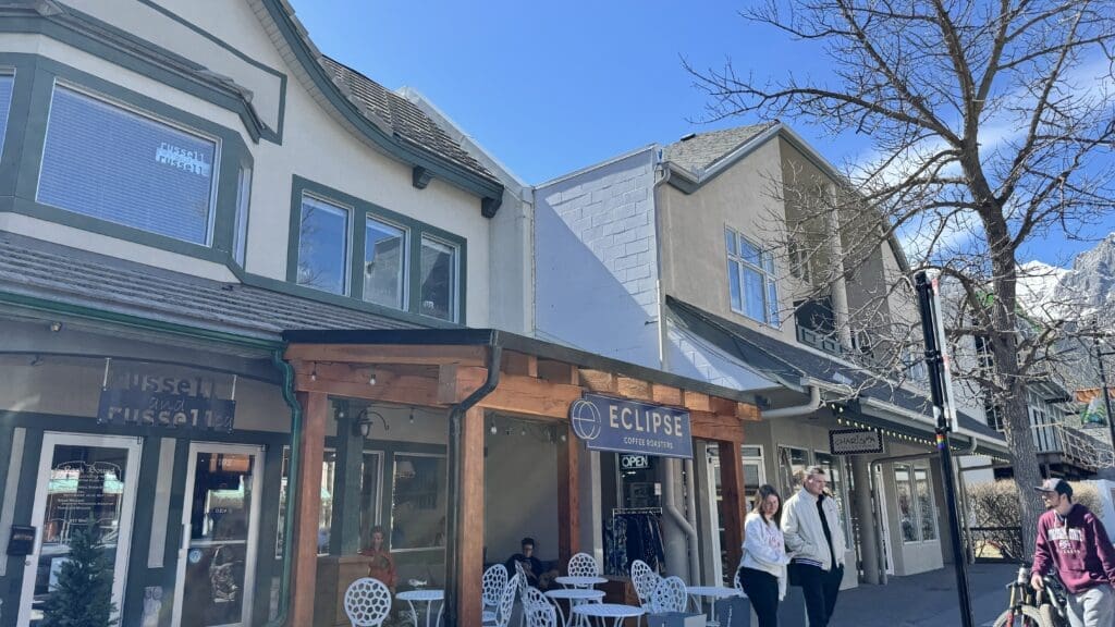 Front view of Eclipse Coffee Roasters in downtown Canmore, nestled in a cozy building with outdoor seating, wooden patio accents, and a scenic mountain backdrop under a clear blue sky. The best Restaurants to Dine in Canmore
