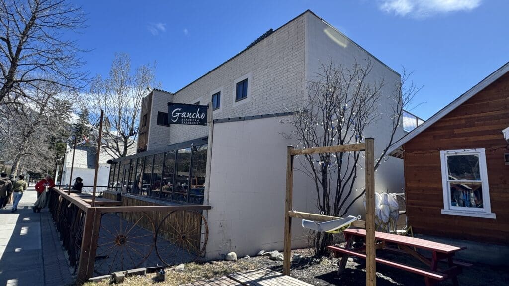 Exterior view of Gaucho Brazilian Barbecue restaurant in Canmore, Alberta, featuring a rustic patio with wagon wheel decor and a white brick facade under a clear blue sky.