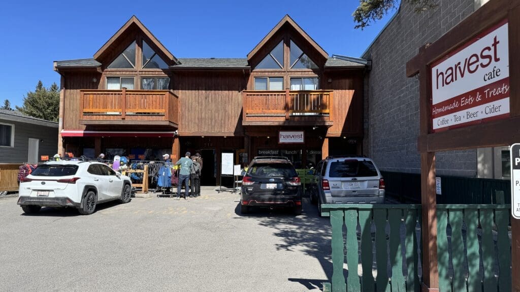 Front view of Harvest Cafe in Canmore, Alberta, showcasing its rustic wooden exterior, outdoor shoppers browsing clothing racks, and a welcoming sign offering coffee, tea, beer, cider, and homemade eats and treats under clear blue skies. The best Restaurants to Dine in Canmore