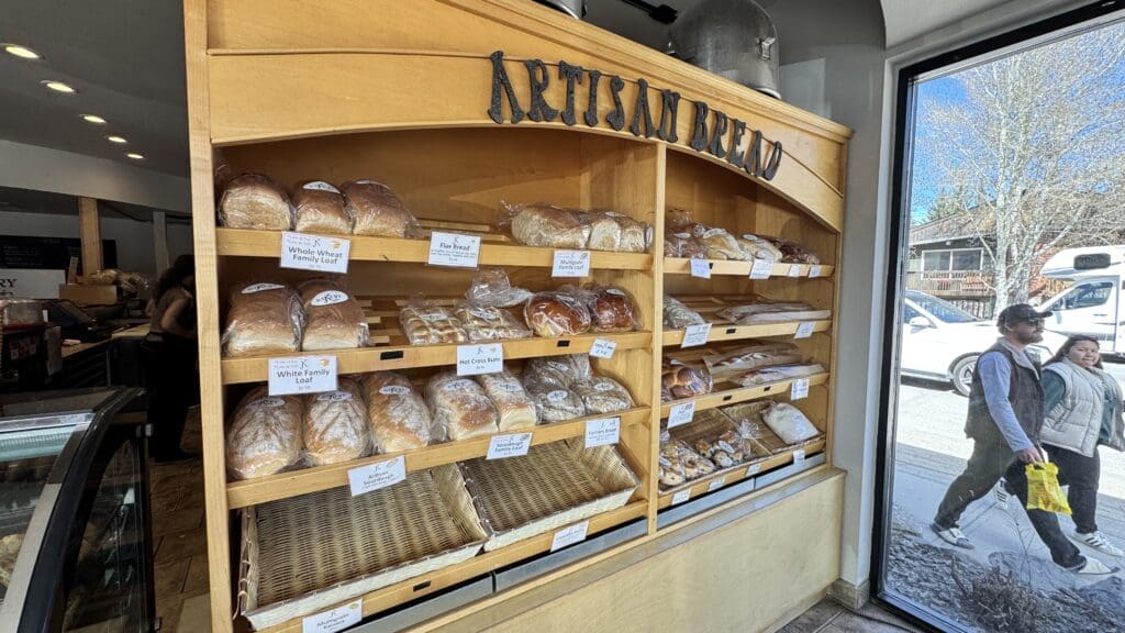 Display shelf of freshly baked artisan breads at JK Bakery Cafe in downtown Canmore, with loaves like sourdough, multigrain, and hot cross buns, viewed from inside the bakery with a street view outside.