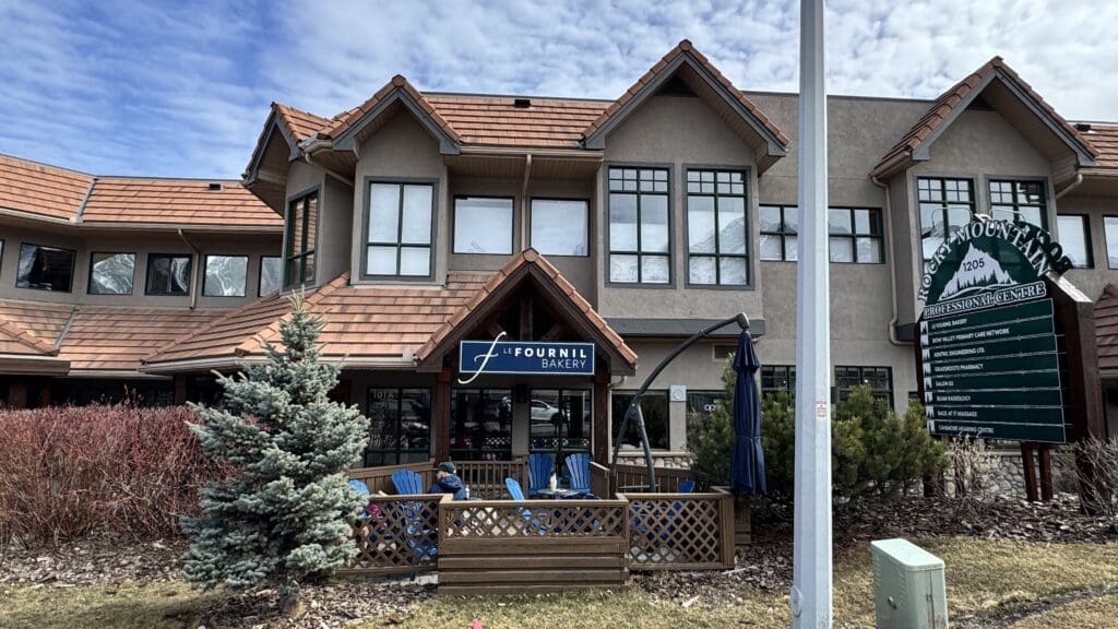 Front facade of Le Fournil Bakery in Canmore, located in the Rocky Mountain Professional Centre, featuring a cozy patio with blue Adirondack chairs and a charming alpine-style roof.