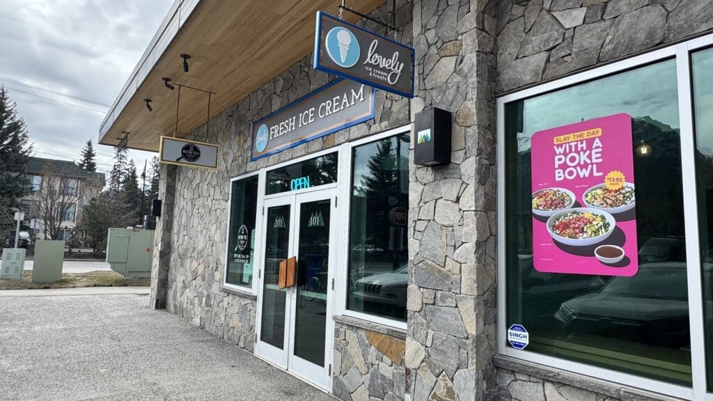 Exterior view of Lovely Ice Cream & Treats in Canmore, Alberta, featuring a stone facade, vibrant signage for fresh ice cream, and a colorful poke bowl advertisement on the window.