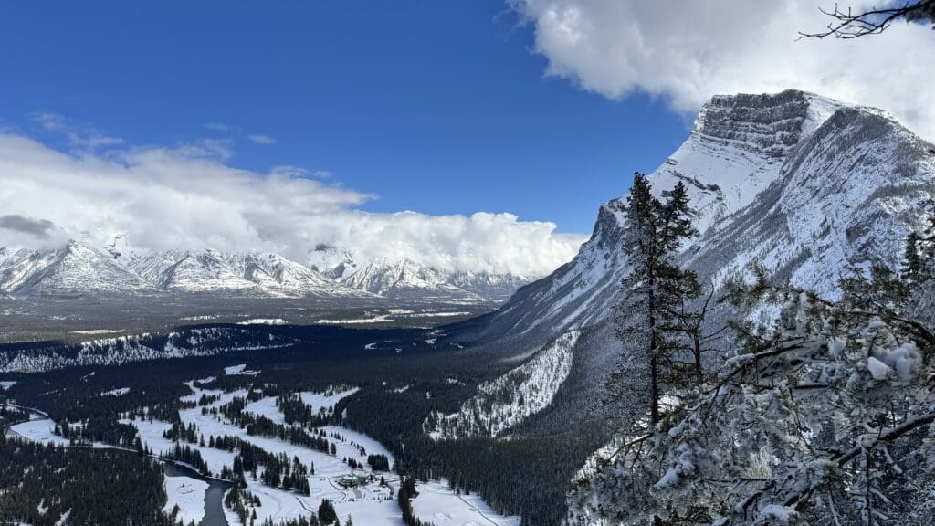 Panoramic winter view from mid-way up Tunnel Mountain Trail in Banff, showcasing the winding Bow River, snow-covered forest, and the towering face of Mount Rundle under a partly cloudy blue sky.