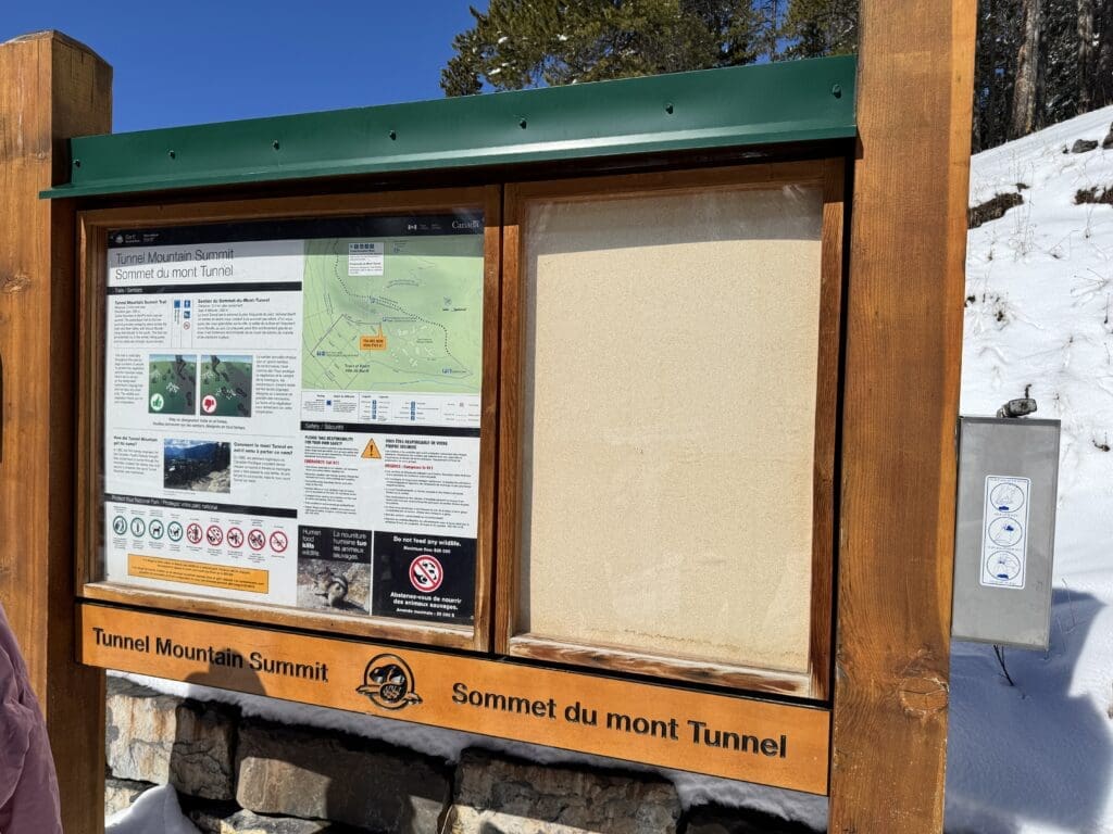 Information board at the Lower Tunnel Mountain Trailhead in Banff National Park, Alberta, showing trail map, safety guidelines, and summit route details under a clear blue winter sky.