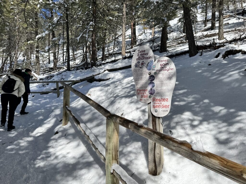 Bilingual Parks Canada sign on Tunnel Mountain Trail in Banff National Park, encouraging hikers to stay on the designated trail and hike responsibly, surrounded by snow-covered trees and a wooden railing in winter.