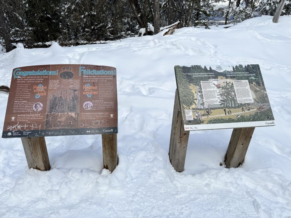 Bilingual Parks Canada summit signs at Tunnel Mountain's peak in Banff National Park, surrounded by fresh snow, celebrating the 1,600-meter elevation achievement and encouraging hikers to stay on the trail.