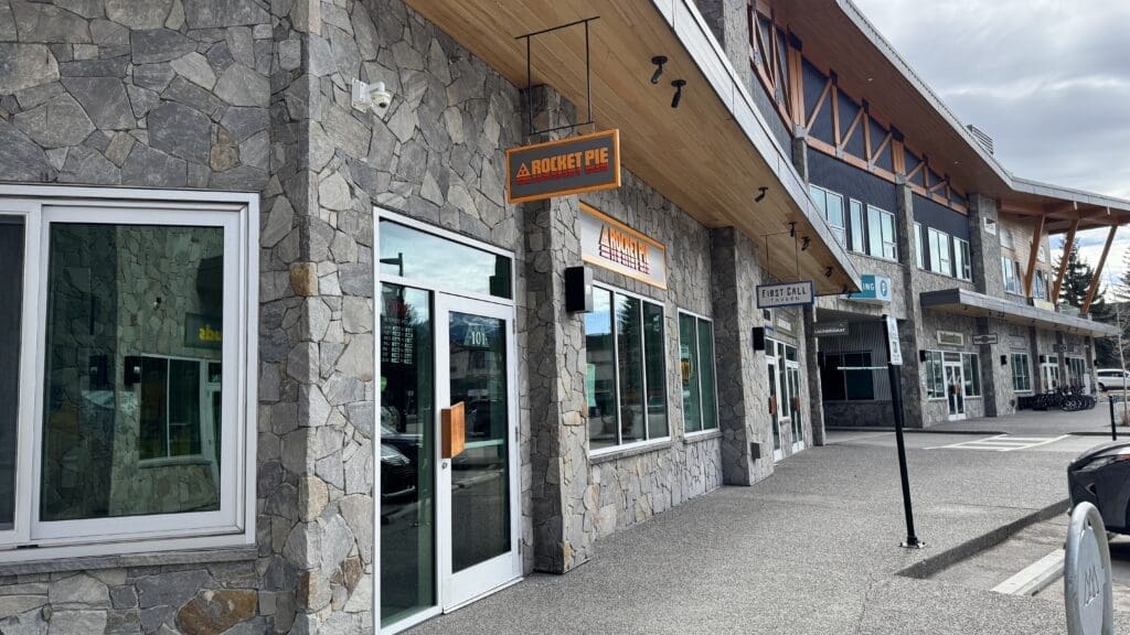 Exterior view of Rocket Pie Pizza in Canmore, Alberta, located in a modern plaza with stone-clad walls, large windows, and a hanging orange sign above the entrance.