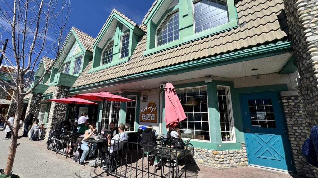 Outdoor seating at Rocky Mountain Bagel Company in Canmore, Alberta, with customers enjoying fresh bagels and coffee under red umbrellas on a sunny day