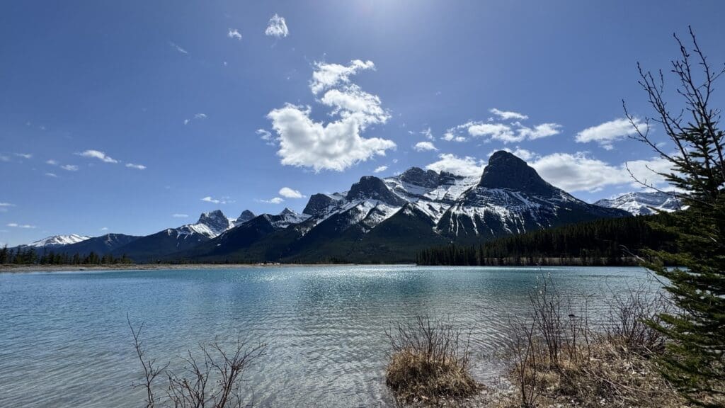 Crystal-clear waters of Rundle Forebay Reservoir reflecting the snow-capped peaks of the Three Sisters mountains in Canmore, Alberta, under a bright blue sky.