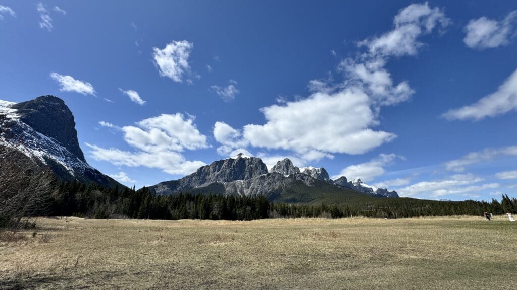 Breathtaking view of the Rocky Mountains near Quarry Lake in Canmore, Alberta, featuring snow-capped peaks, a wide open meadow, and a blue sky dotted with fluffy white clouds.