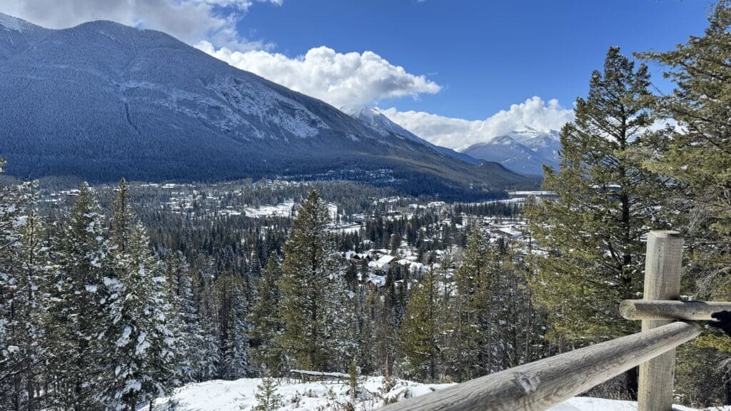 Scenic viewpoint along the lower Tunnel Mountain Trail in Banff, Alberta, with a wooden railing in the foreground overlooking a snow-covered forest and the town of Banff nestled against the backdrop of towering, rugged mountains under a bright blue winter sky.