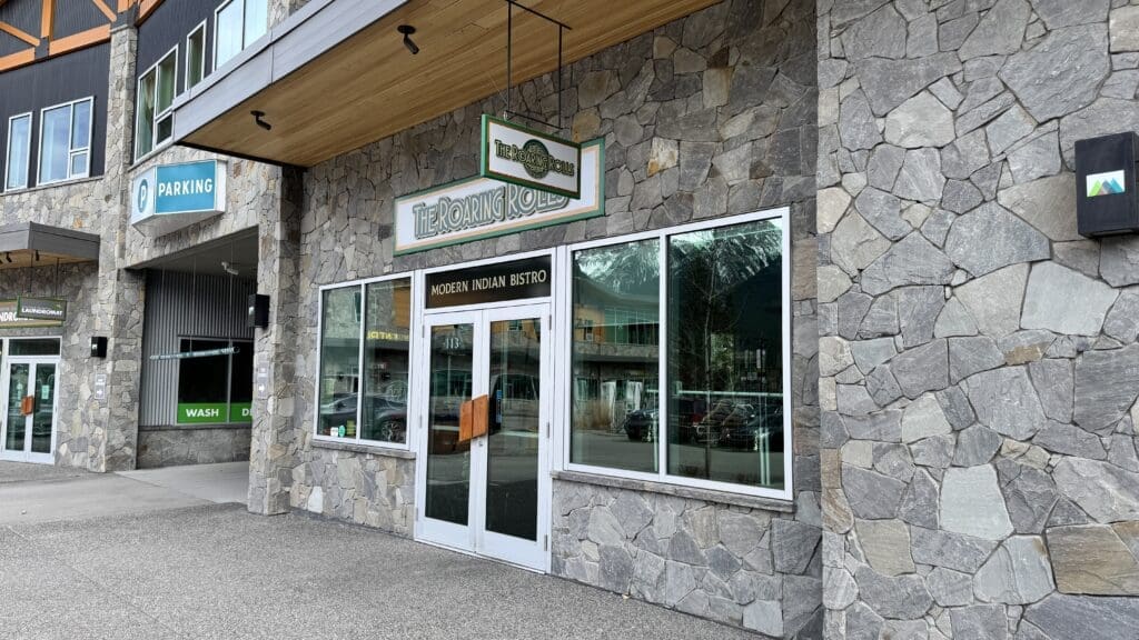 Exterior view of The Roaring Rolls, a modern Indian bistro in Canmore, Alberta, featuring a stone facade and large glass windows with mountain reflections.