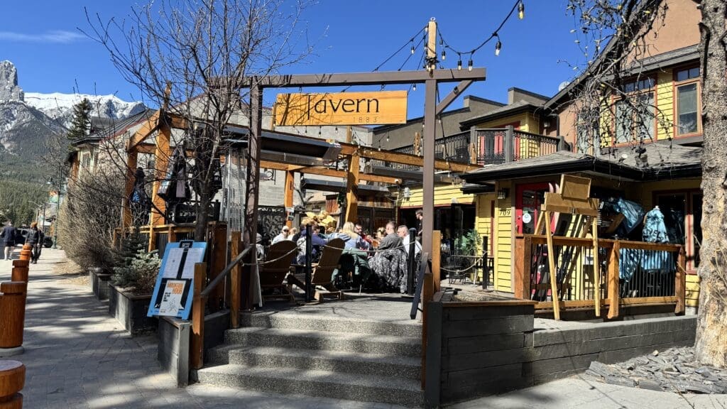 Guests enjoy a sunny afternoon on the outdoor patio at Tavern 1883 in Canmore, Alberta, surrounded by cozy wooden beams, colorful architecture, and a backdrop of the Rocky Mountains.