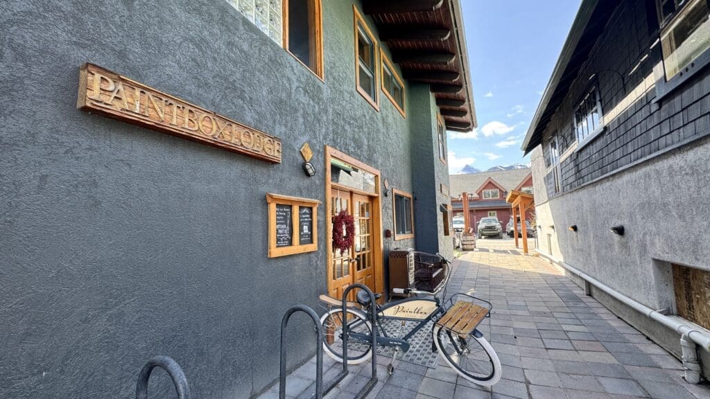 Front entrance of The Paintbox Lodge in Canmore, Alberta, featuring rustic wooden signage, a welcoming wreath on the door, and a branded bicycle parked outside on a charming stone pathway.