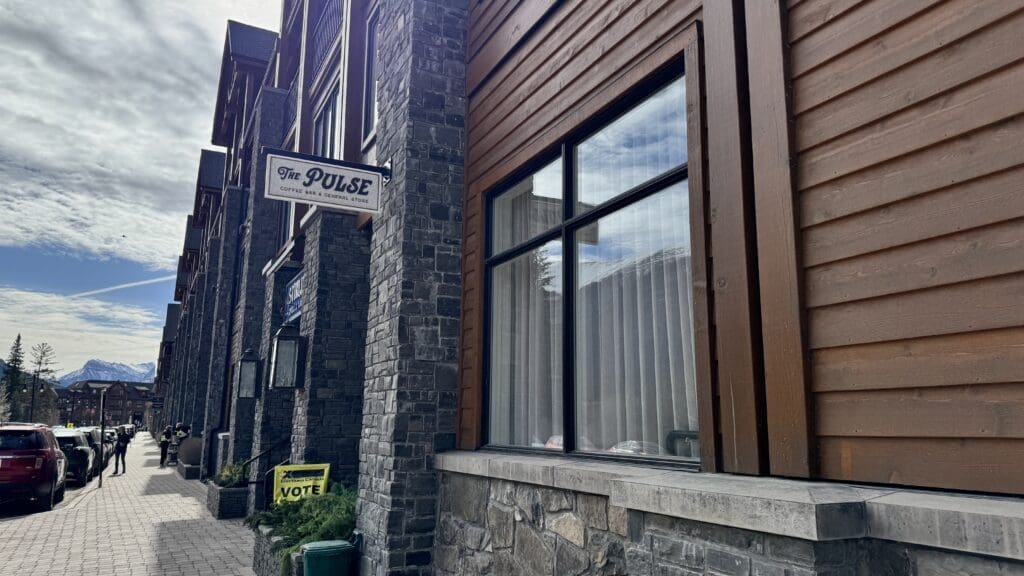 Street view of The Pulse General Store + Coffee Bar in Canmore, Alberta, featuring rustic stone and wood exterior, nestled along a scenic mountain town sidewalk with views of the Rockies in the background. The best Restaurants to Dine in Canmore