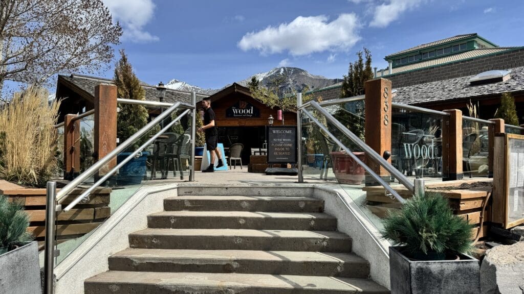 The Wood Restaurant & Lounge in Canmore, Alberta, with a rustic log cabin design, large outdoor patio, and stunning Rocky Mountain views under a bright blue sky.