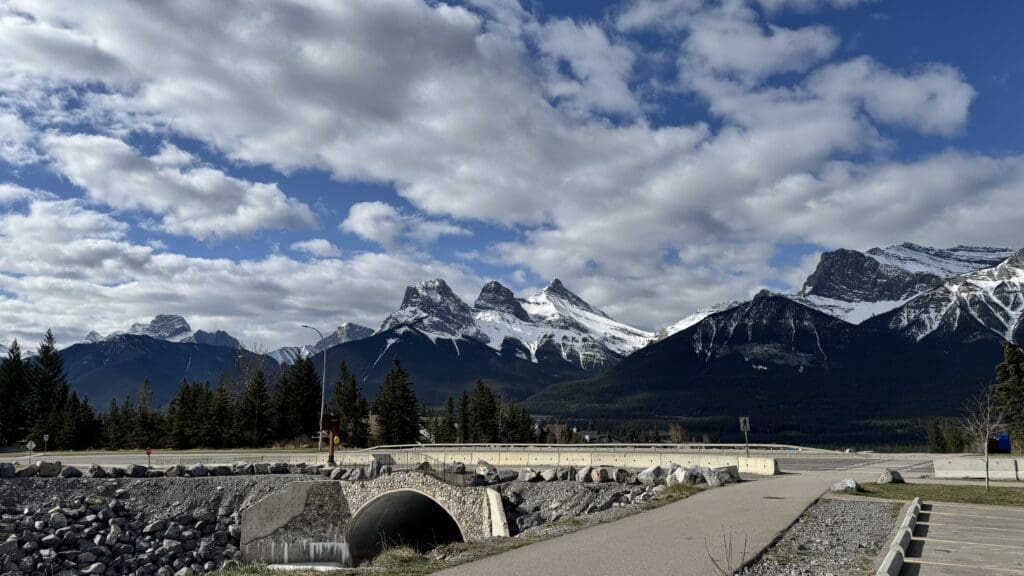 Scenic view of the iconic Three Sisters peaks from the Cougar Creek Trailhead in Canmore, Alberta, with a pedestrian bridge and cloudy sky enhancing the dramatic mountain backdrop.