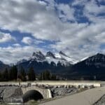 Scenic view of the iconic Three Sisters peaks from the Cougar Creek Trailhead in Canmore, Alberta, with a pedestrian bridge and cloudy sky enhancing the dramatic mountain backdrop.