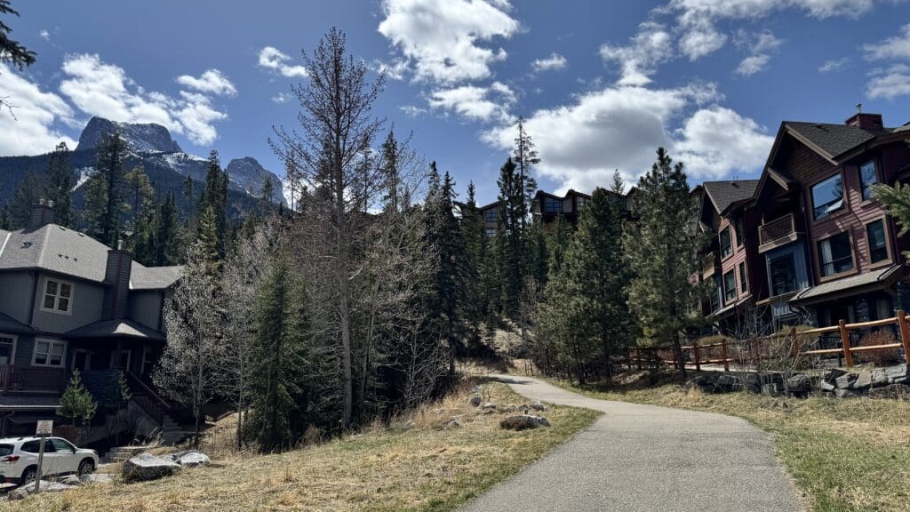 Scenic view of the Three Sisters Pathway in Canmore, Alberta, with modern mountain homes on one side, pine forest on the other, and the majestic snow-capped Three Sisters peaks in the background under a bright blue sky.