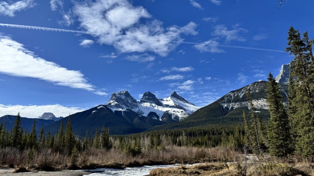 Stunning view of the Three Sisters mountain peaks near Canmore, Alberta, under a vivid blue sky with forested foreground and a partially frozen stream