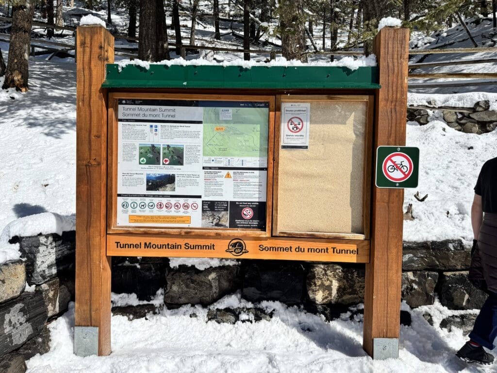 Trailhead signboard at the Upper Tunnel Mountain Trail in Banff National Park, Alberta, surrounded by snow and evergreen trees, providing hikers with safety information, trail maps, and a clear “no bicycles” symbol for winter trail use.