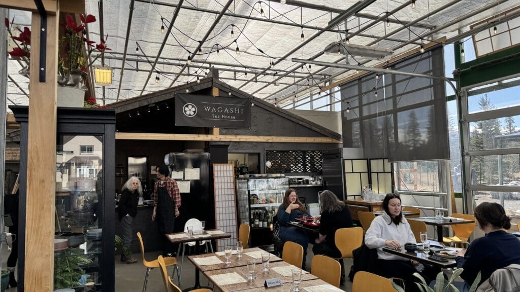 Interior of Wagashi Tea House in Canmore, Alberta, featuring a cozy greenhouse-style setting with minimalist Japanese décor, wooden furniture, string lights, and guests enjoying traditional Japanese tea and cuisine.