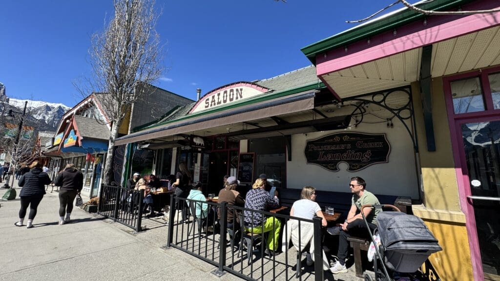 Outdoor patio seating at Where The Buffalo Roam Saloon in Canmore, Alberta, with patrons enjoying food and drinks under clear blue skies along a lively mountain town street.