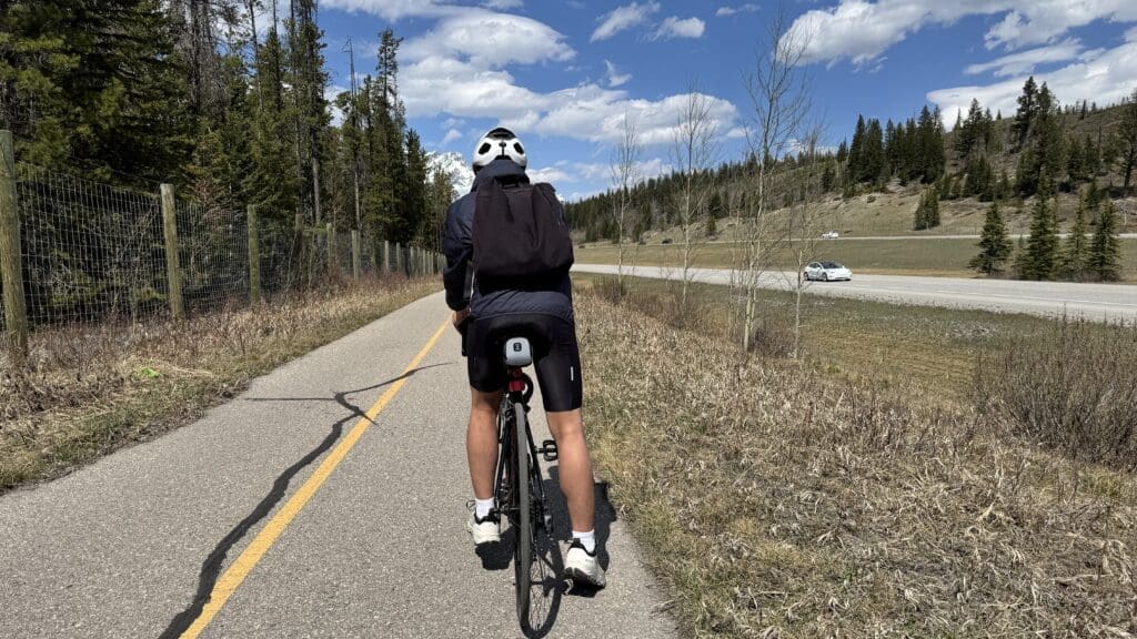 Biker rides on a sunny Legacy Trail in Banff National Park lined with trees and grass. A mountain and a car on a road are visible in the distance under a blue sky with clouds
