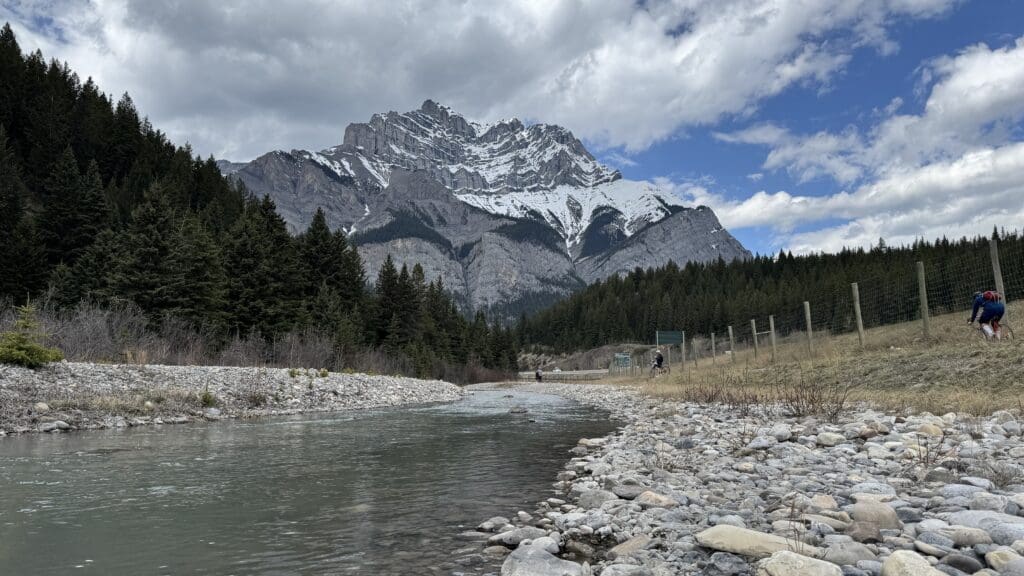 Snow-capped Cascade Mountain under cloudy sky, pine forest below. A calm Bow River with rocky banks in foreground; cyclist rides on Legacy Trail in Banff National Park