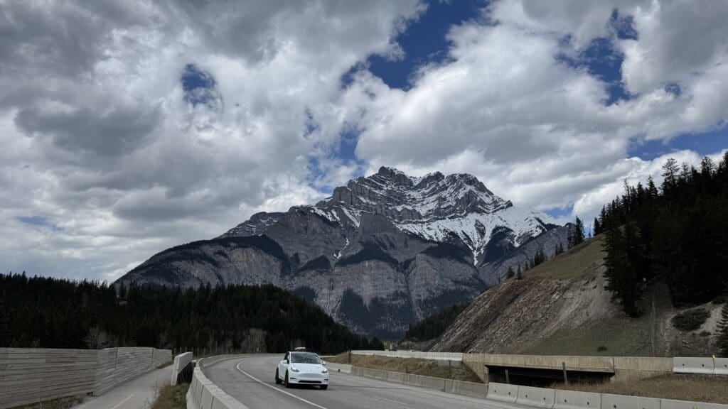 A white car drives along a curved mountain highway in Banff National Park under a dramatic sky with scattered clouds. Snow-capped Cascade Mountain and dense forests create a majestic backdrop.