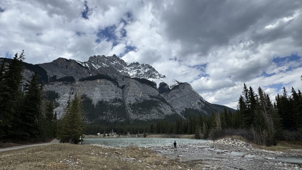 Cascade Mountain landscape with a cloudy sky, snowy peaks, and a flowing Cascade Ponds in Banff National Park. Pine trees line the shores, and a person stands by the water, evoking serenity.