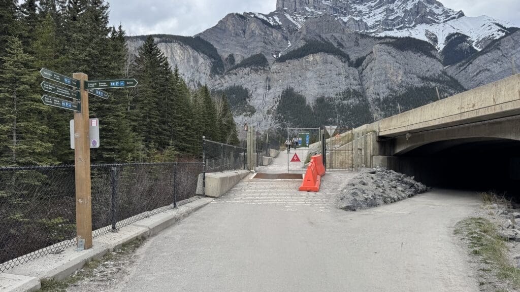 Pathway with a wooden signpost on Legacy Trail in Banff National Park showing hiking trails, bordered by fencing. Behind, a majestic mountain range looms under a cloudy sky, conveying a sense of adventure.