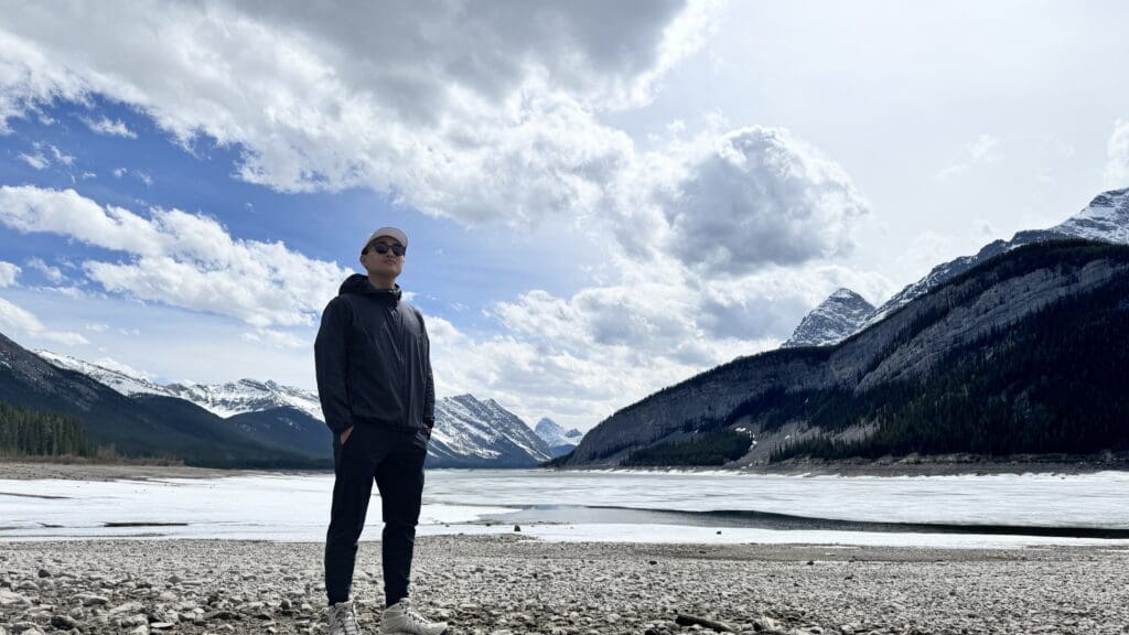 A person in sunglasses and a black jacket stands on a rocky shore by a partially frozen Spray Lakes in Kananaskis, Alberta. Snow-capped mountains and a cloudy sky are in the background.
