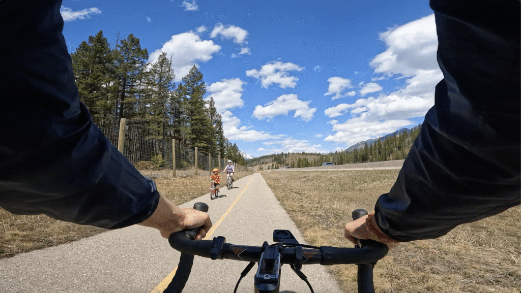 Cyclist view of Legacy Trail in Banff National Park through a forest; two people biking ahead. Clear blue sky with clouds, creating a serene outdoor atmosphere.